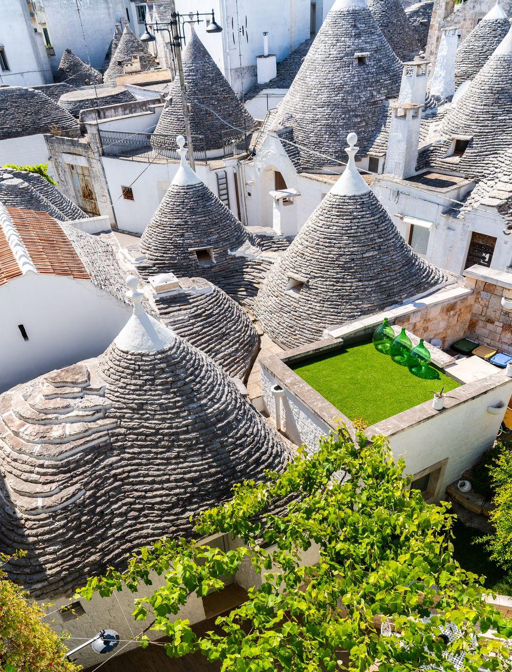 I tetti a cono dei trulli di Alberobello visti dall’alto, tra case bianche, una terrazza con tre damigiane verdi e i rami di una vite