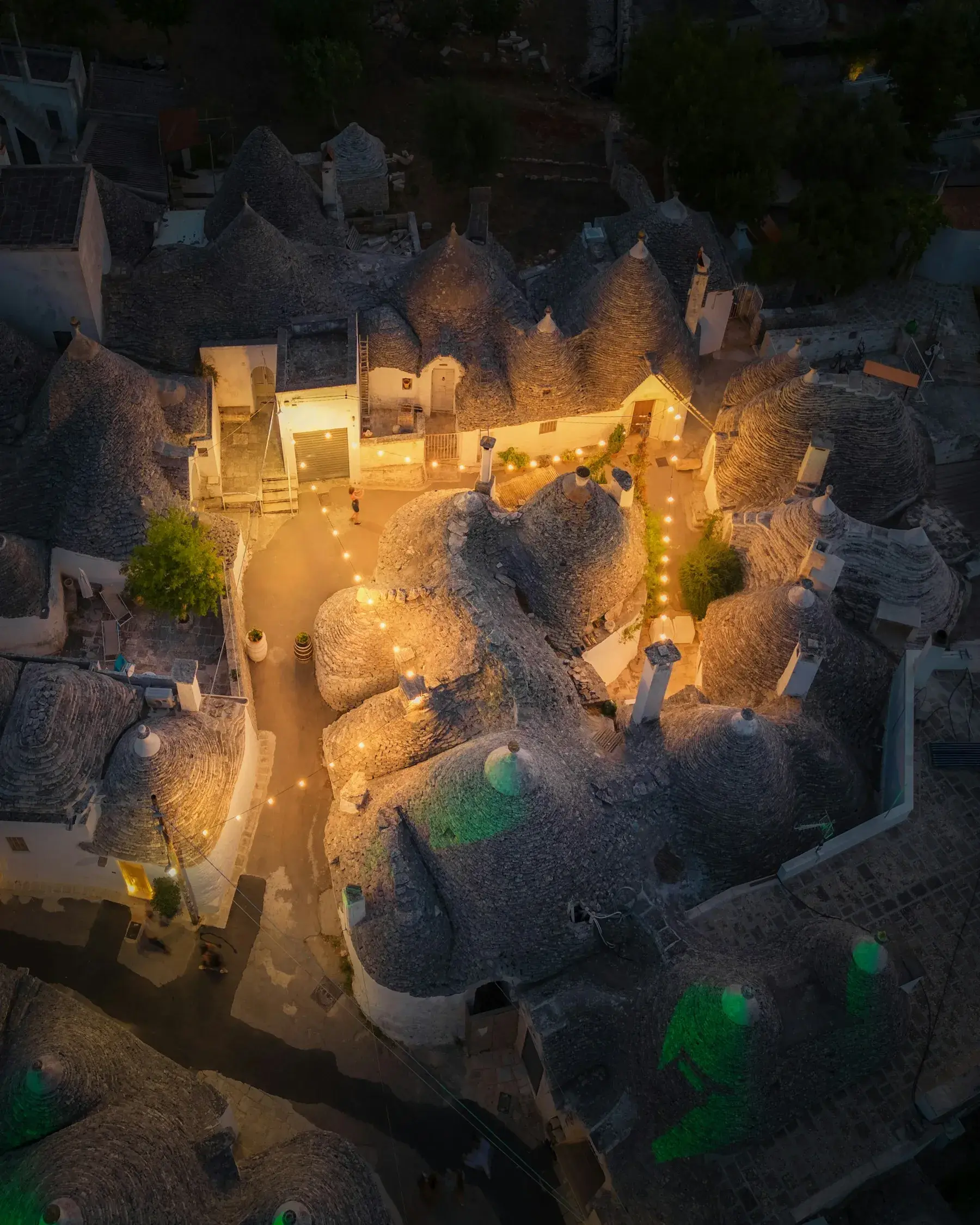Coberturas de trulli vistas de cima ao entardecer, com fios de luzes quentes entre as casas. Alberobello.