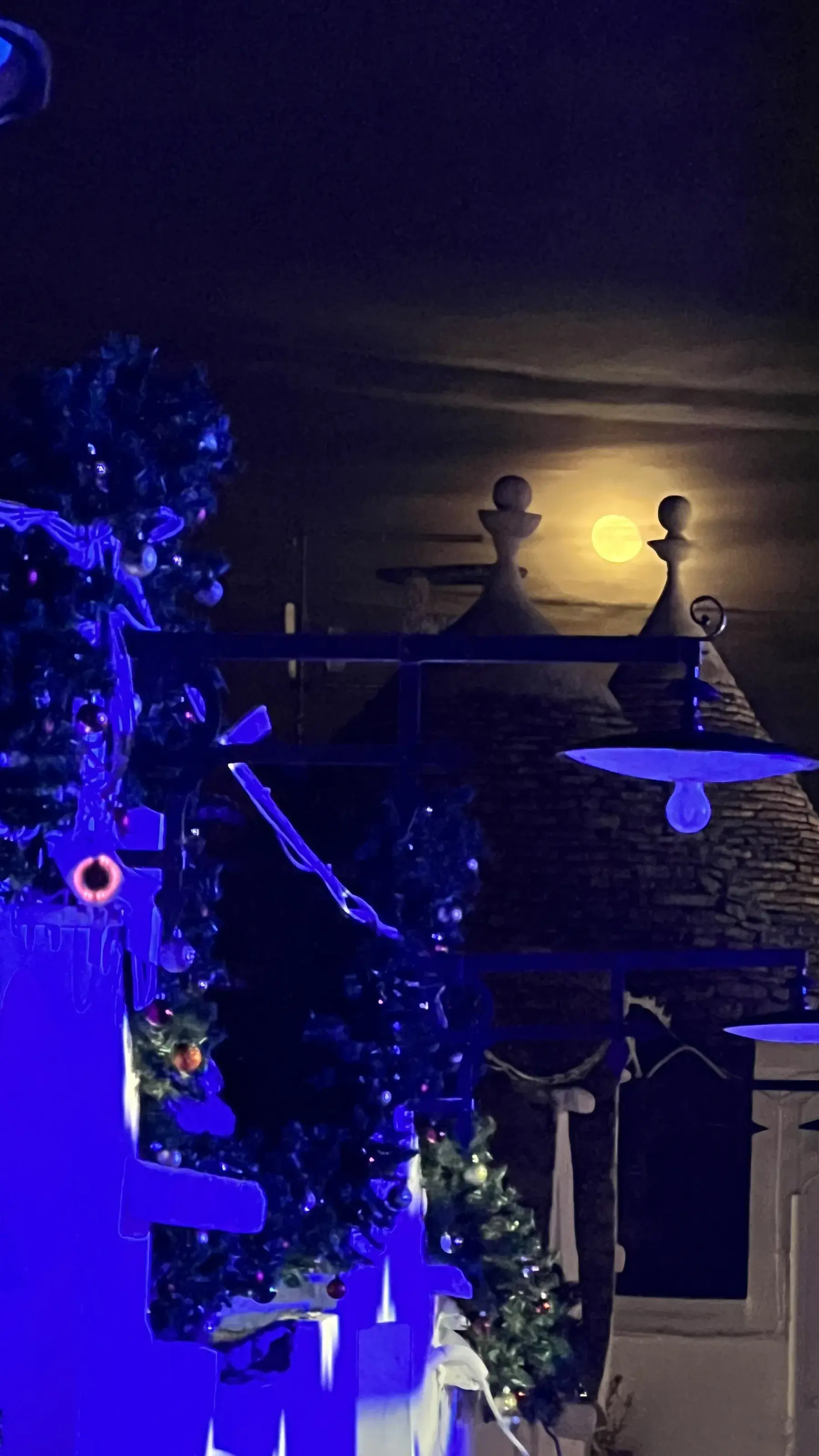 Uma guirlanda e uma luminária azul diante de coberturas de trulli à noite, com a lua cheia atrás. Alberobello.