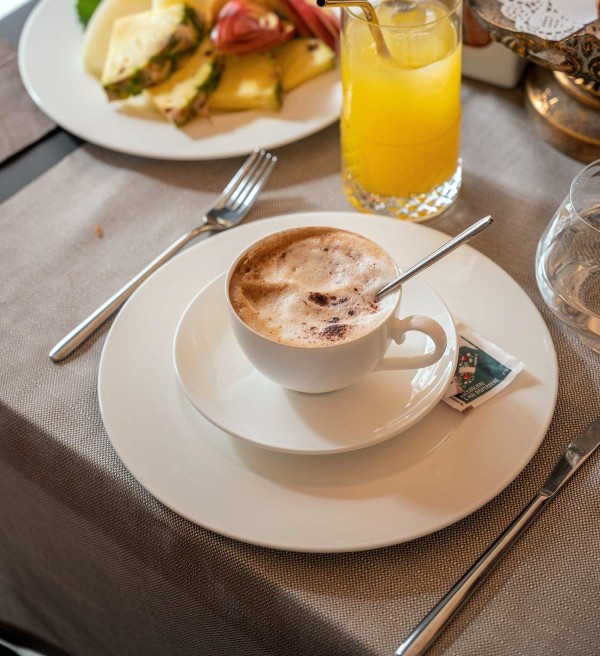 Colazione da NOS: cappuccino con cacao, un succo e un piatto di frutta fresca tagliata