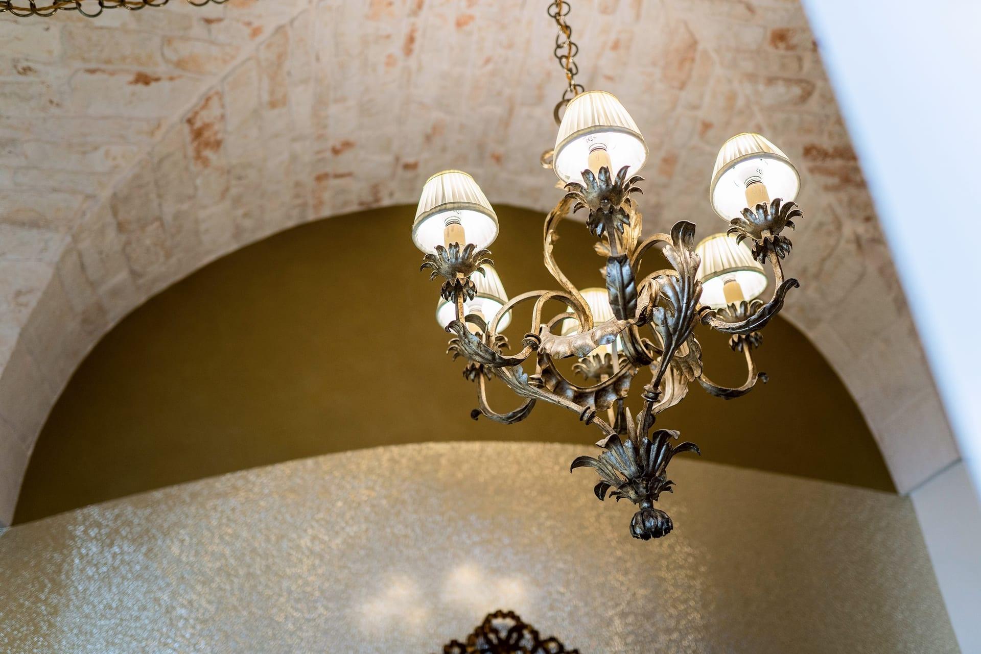 Foresteria — der Metallkronleuchter mit Blattmotiven und plissierten Schirmen, unter dem Steinbogen