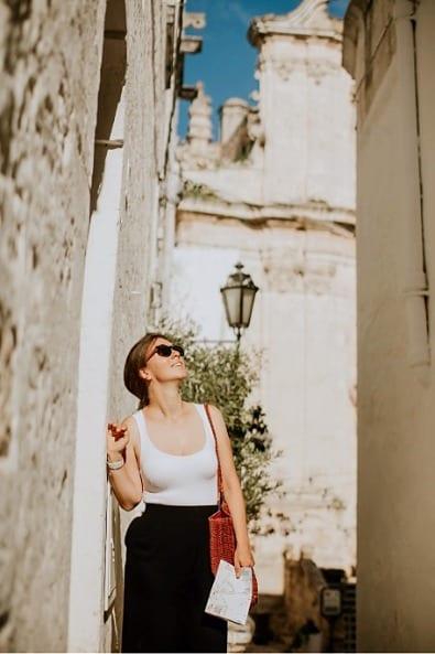 Una donna con una cartina in mano, in un vicolo stretto tra muri bianchi, alza lo sguardo verso una facciata barocca