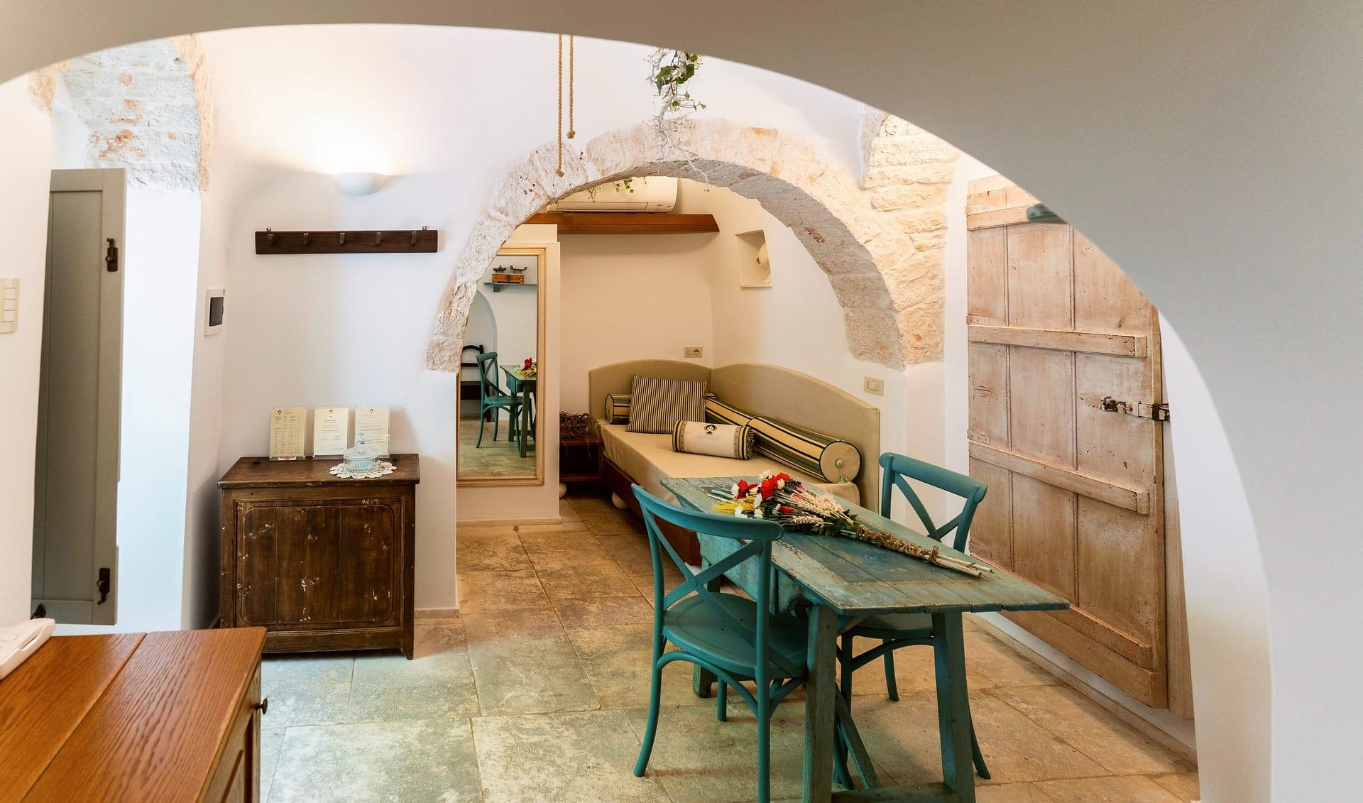 Suite Trullo — durch den weißen Bogen der Tisch mit Blumen und das Sofa mit den gestreiften Nackenrollen