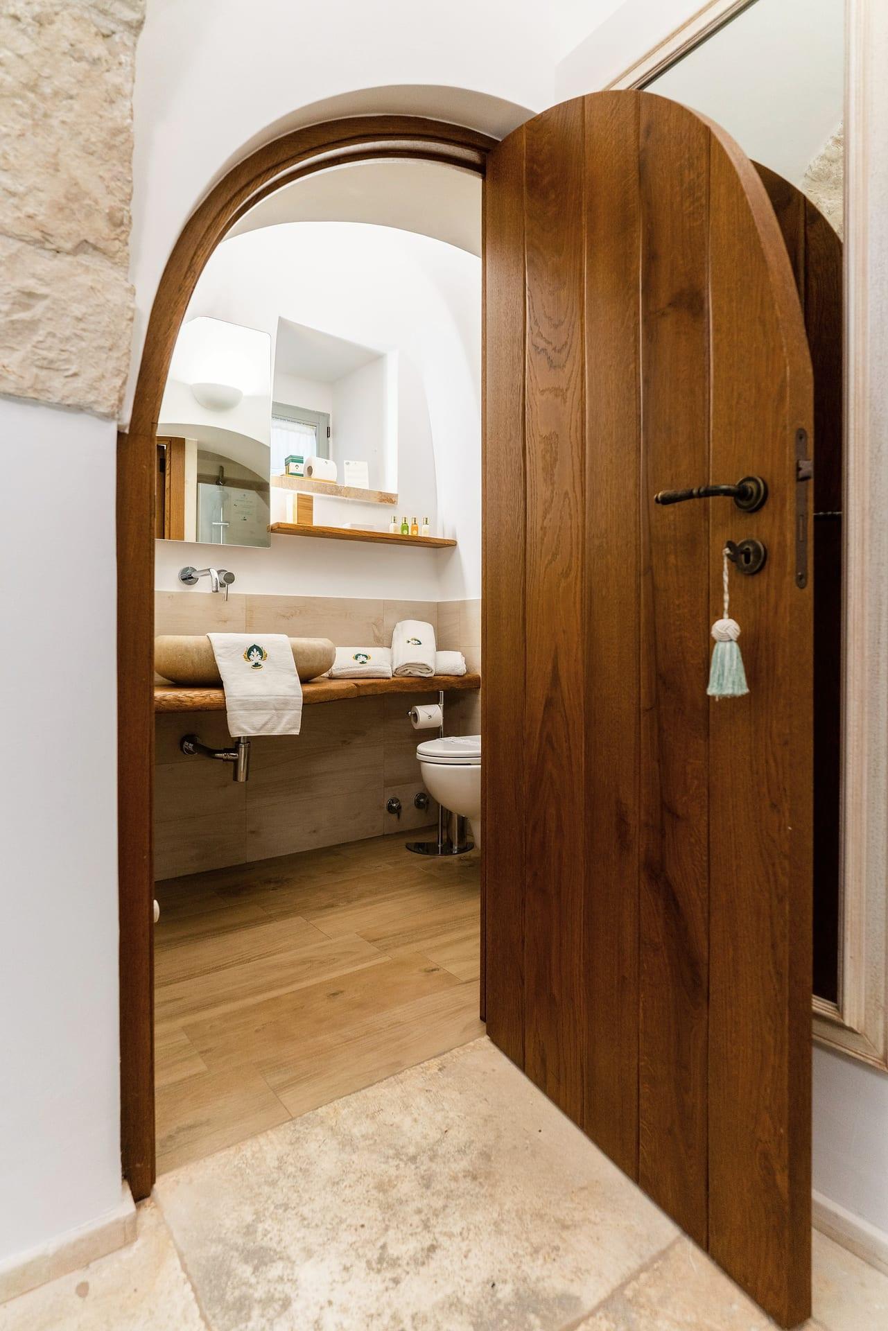Suite Trullo — die hölzerne Bogentür, offen zum Bad, mit der hellblauen Quaste am Schlüssel