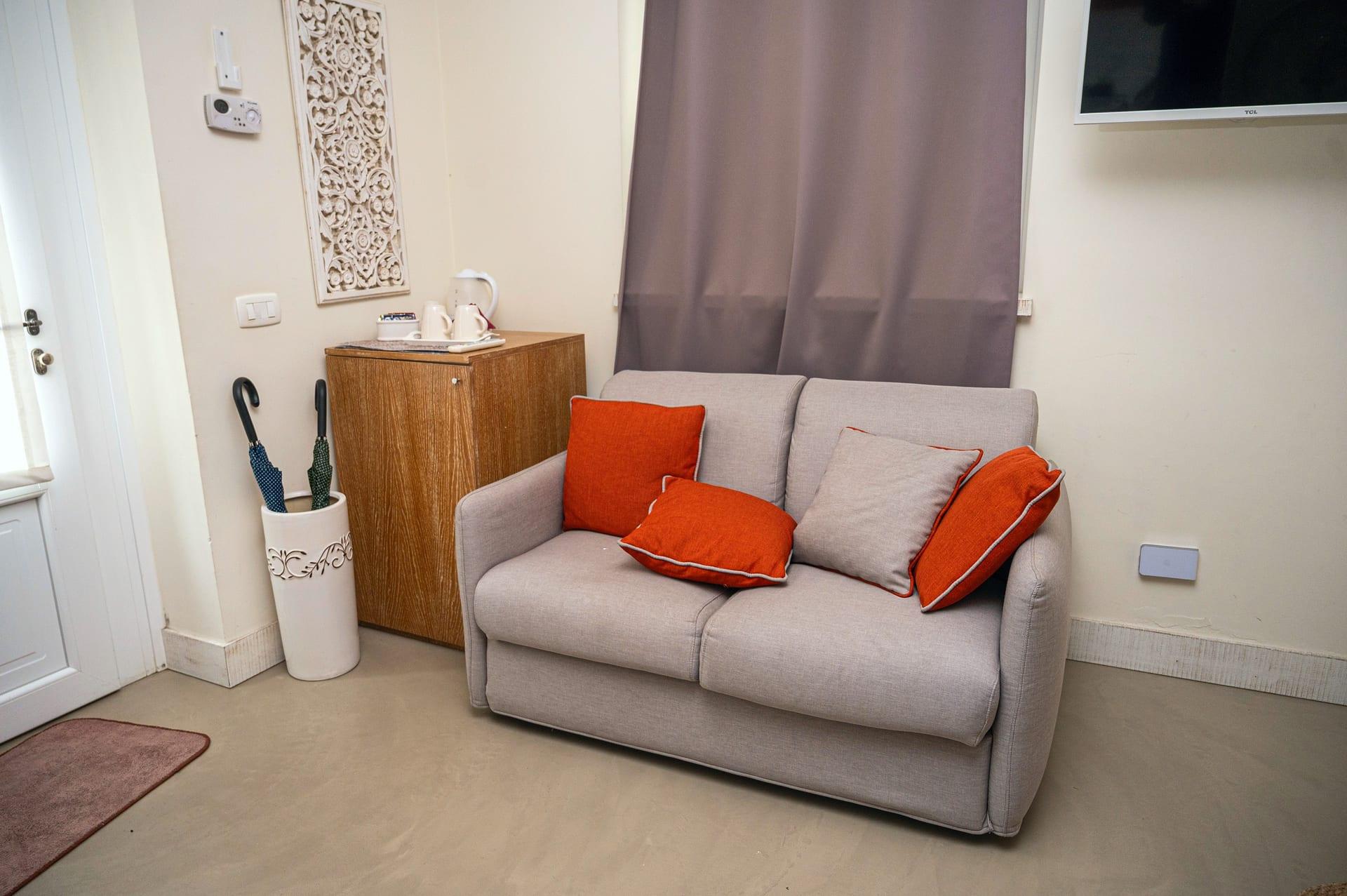 La Piccola — das kleine graue Sofa mit orangefarbenen Kissen, neben dem Schränkchen mit dem Wasserkocher