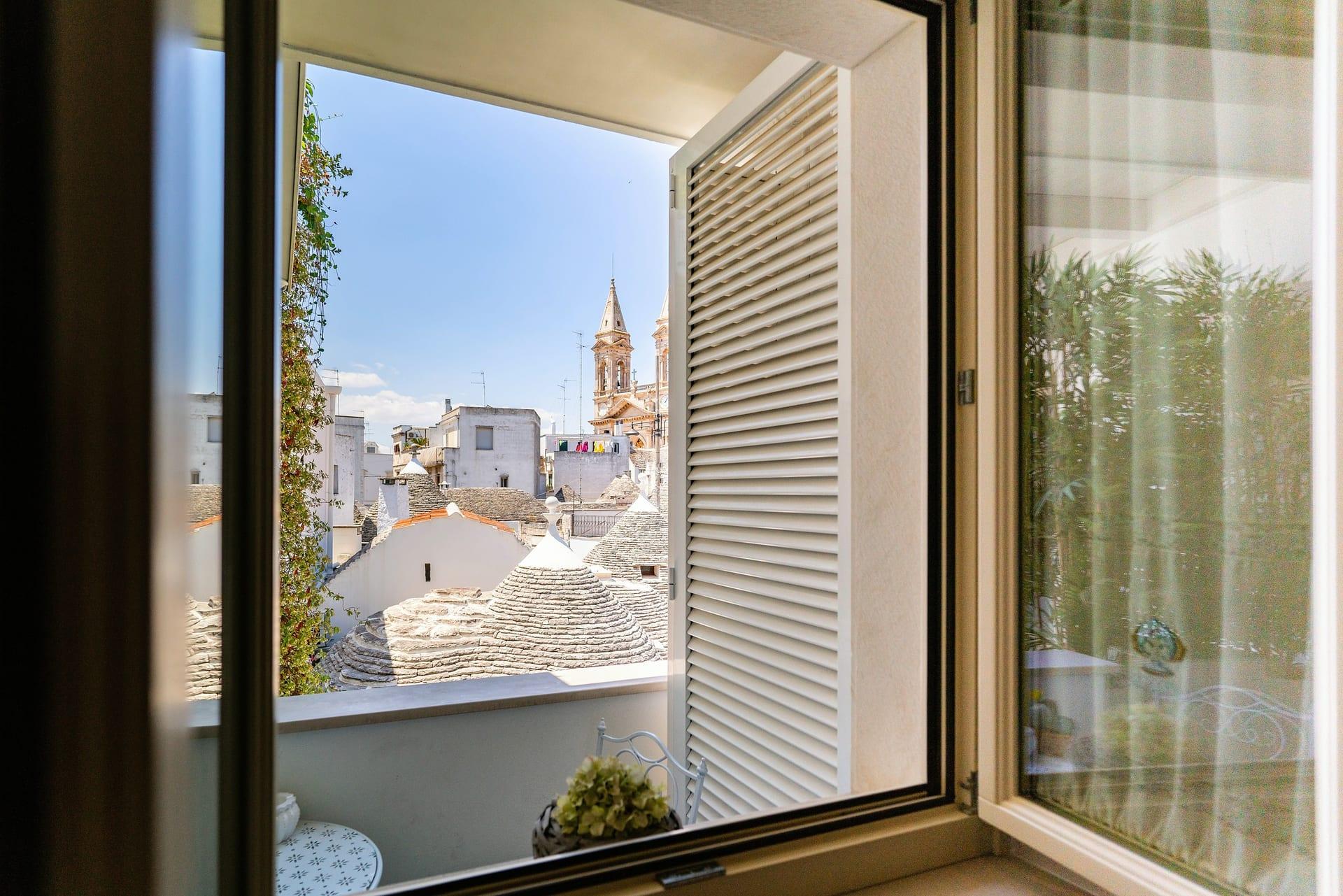 Nobile Suite — das offene Fenster mit weißem Lamellenladen, mit Blick auf die Kegeldächer der Trulli und den Glockenturm