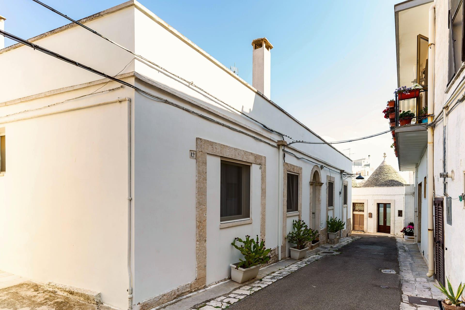 Pietra — la fachada encalada con los marcos de piedra y, al fondo de la calle, un trullo