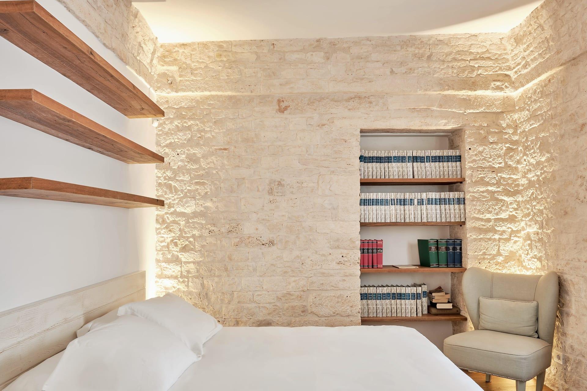 Pietra — la estantería de libros en el nicho de piedra, con el sillón gris junto a la cama