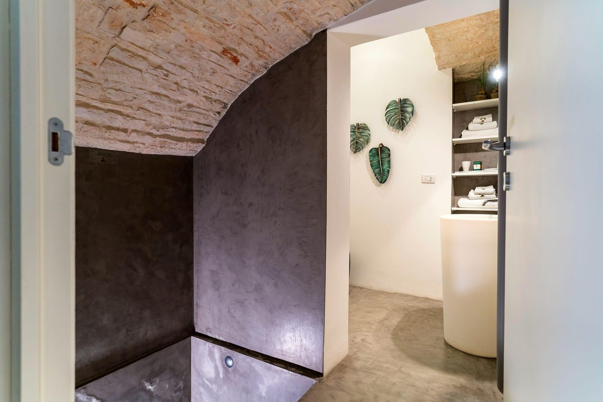 Pietra — el baño con las paredes grises bajo la bóveda de piedra y, tras la puerta, el lavabo de columna