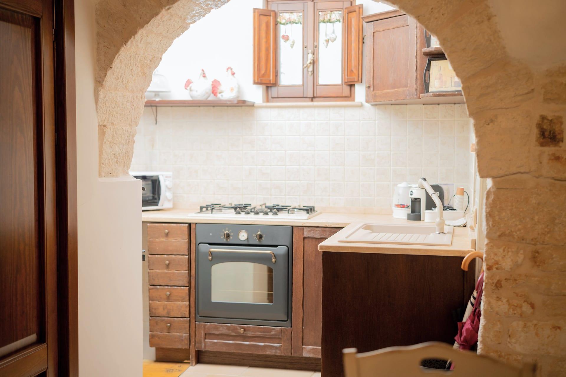 Trullo Maestro — die Küche durch den Steinbogen gesehen, mit dem grauen Backofen und den Keramikhühnern