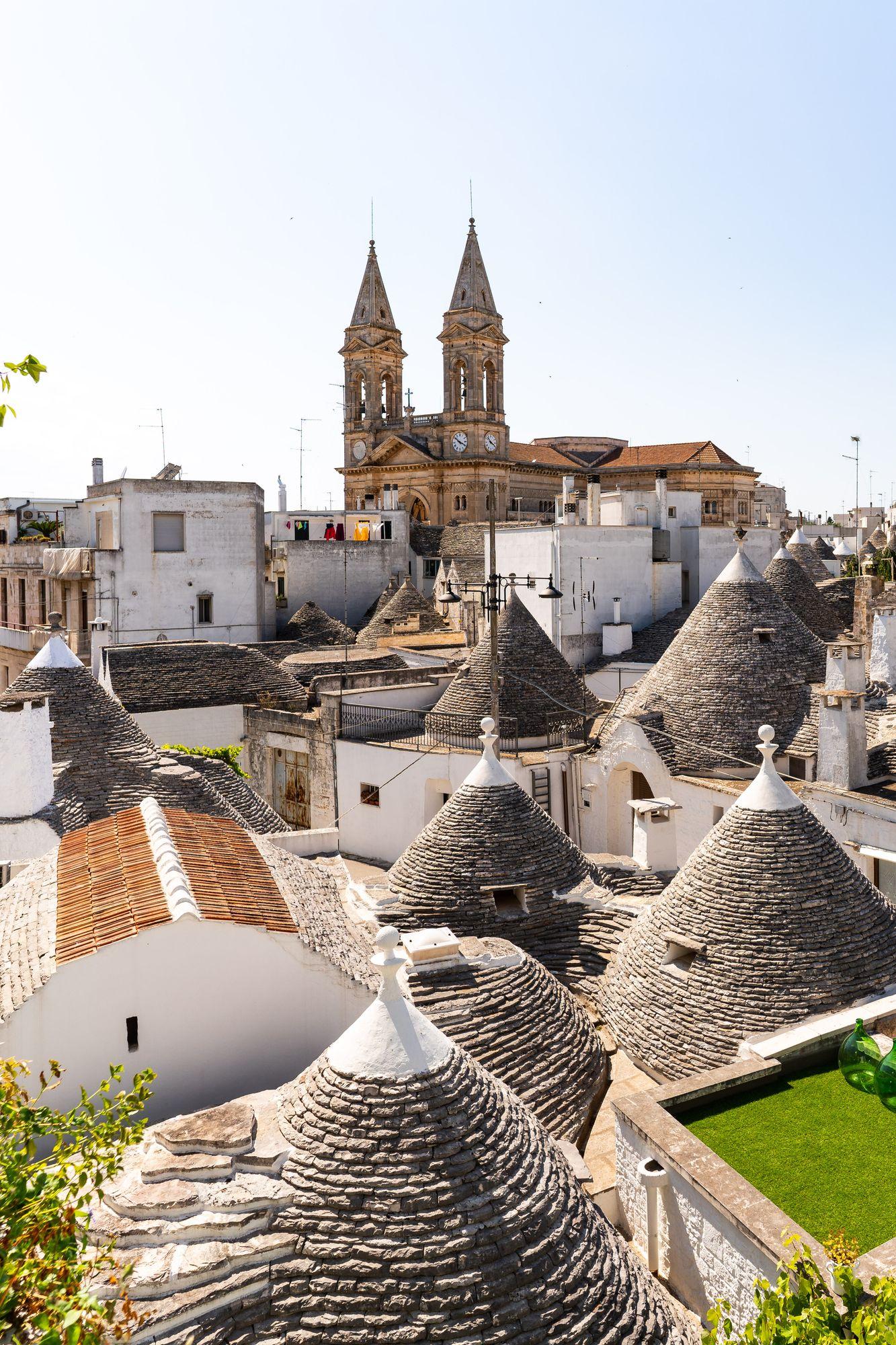 Die Kegeldächer der Trulli und die weißen Häuser von Alberobello von oben, dahinter die Glockentürme der Basilika