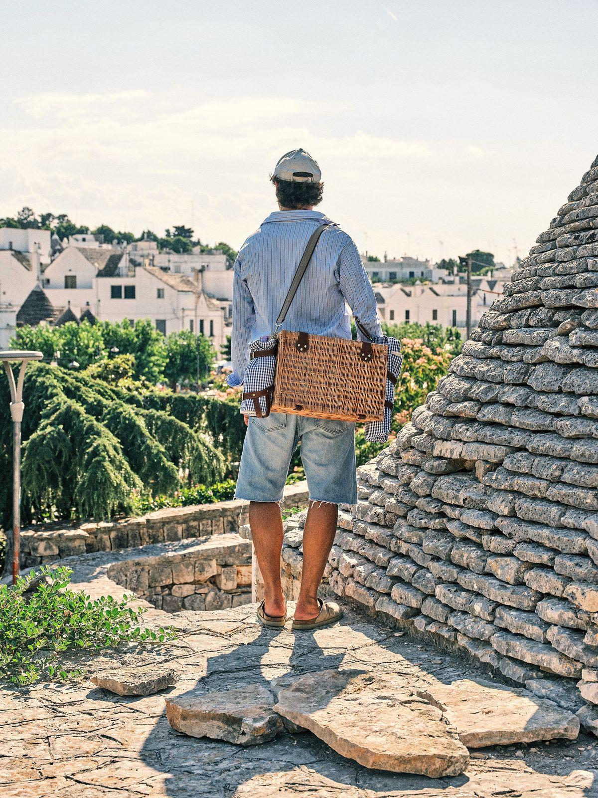 Ein Mann von hinten, einen Picknickkorb über der Schulter, blickt auf die Trulli und die weißen Häuser von Alberobello