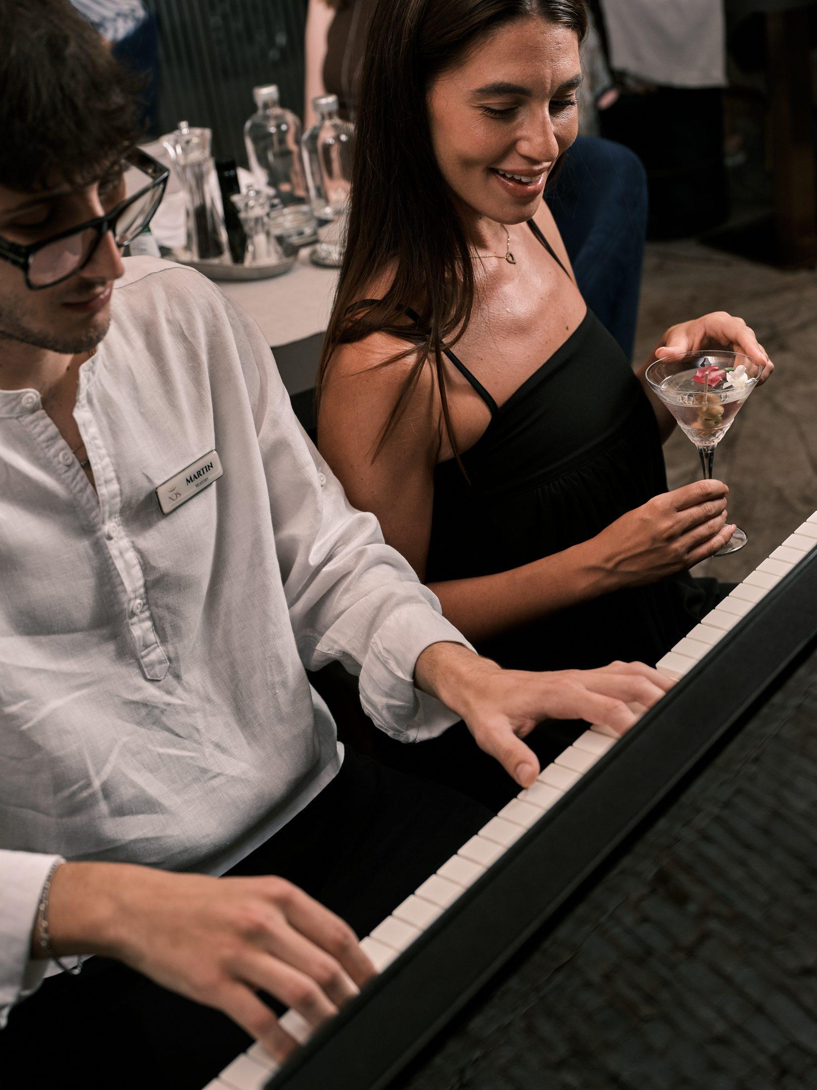 Der Pianist spielt; neben ihm lächelt ein Gast mit einem Glas in der Hand