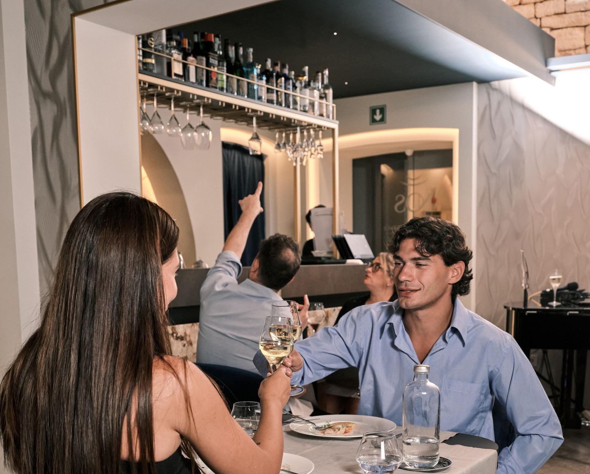Die Bar von NOS mit den Flaschen und den hängenden Gläsern; davor stößt ein Paar am Tisch an