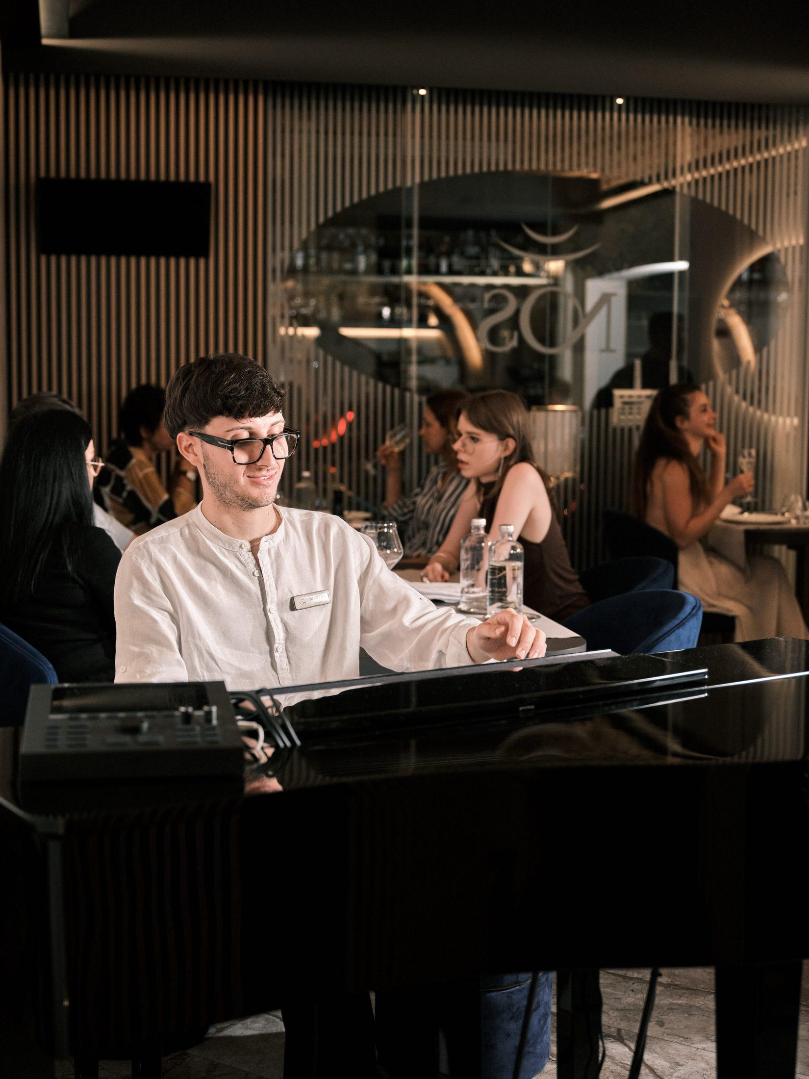 Der Saal von NOS: der Pianist am Klavier; dahinter Gäste an den Tischen und der Schriftzug NOS auf dem Glas
