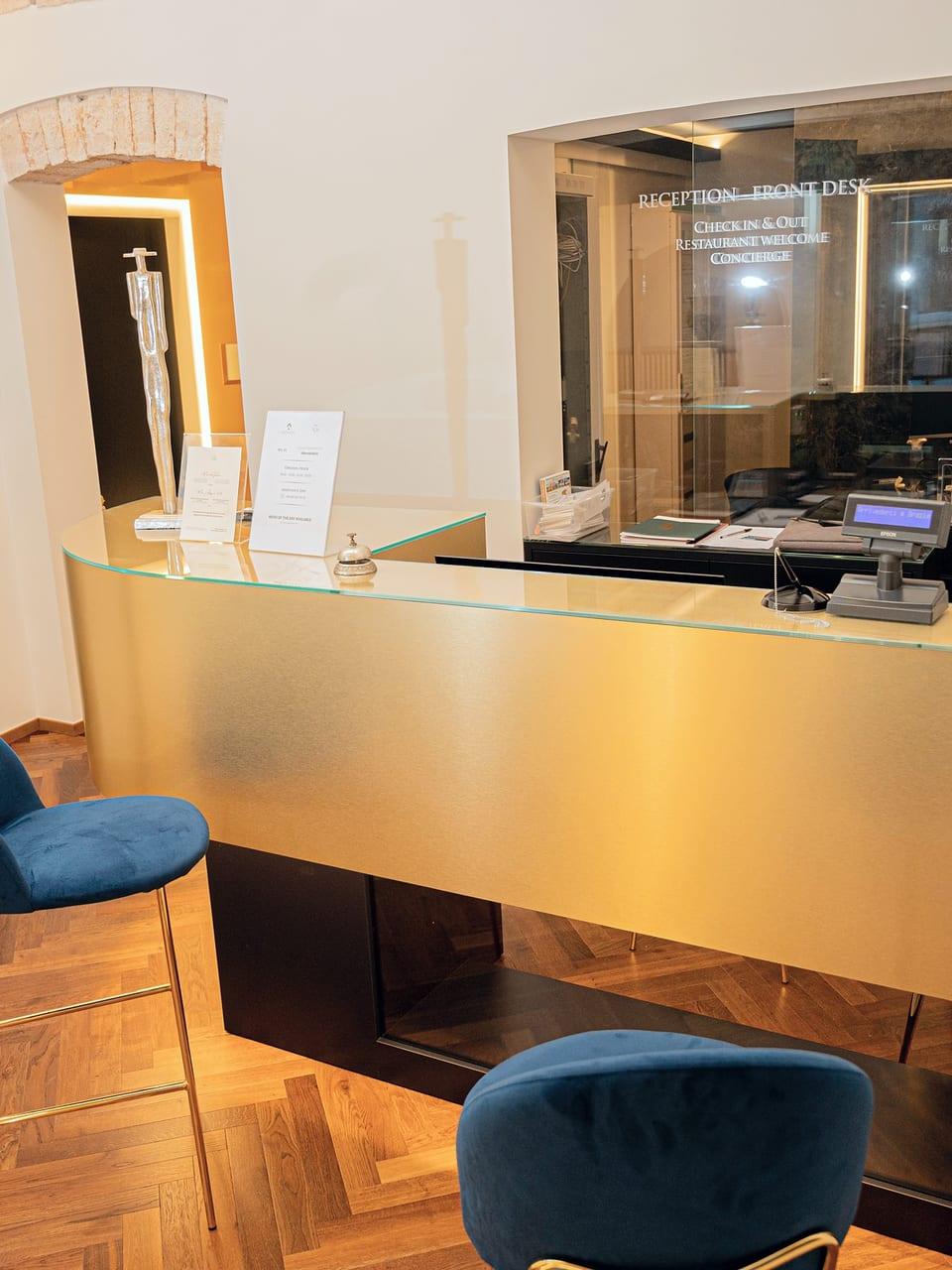 Der Rezeptionstresen aus Messing, mit dem Schriftzug „Reception · Front desk“ auf dem Glas und den blauen Samthockern