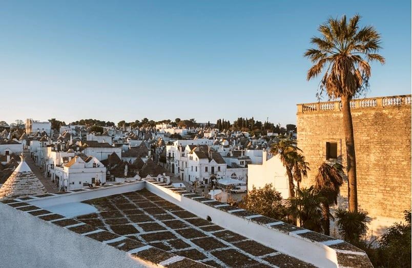 Alberobello von oben im goldenen Licht: die Kegeldächer der Trulli, die weißen Häuser und Palmen neben einem Steinbau