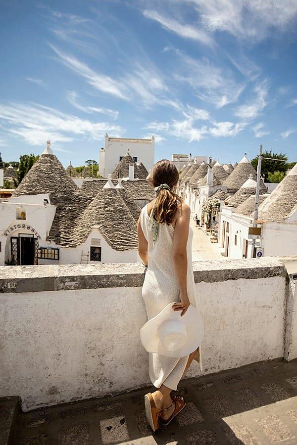Eine Frau, von hinten gesehen, den Hut in der Hand, blickt von oben auf die Kegeldächer der Trulli und die Dorfstraße