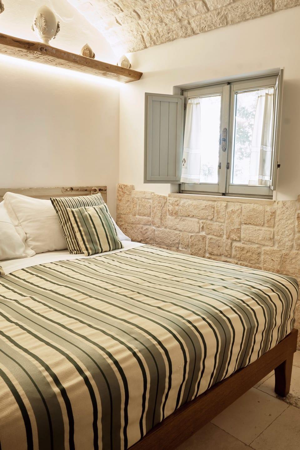 Suite Trullo — das Bett neben der Natursteinwand und dem Fenster mit blauen Läden