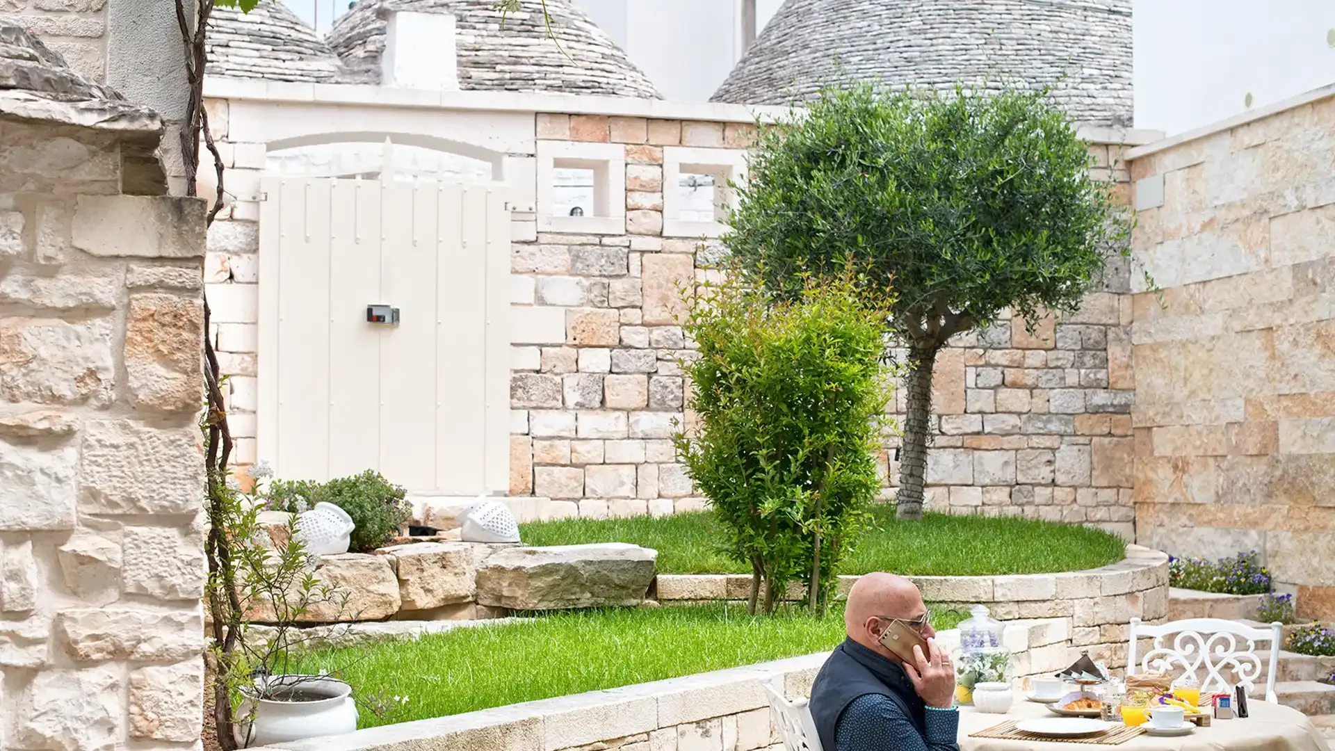Man on phone in a stone courtyard with breakfast on the table.
