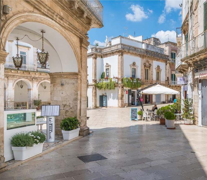 Piazza soleggiata con edifici storici, caffè all'aperto e negozi.