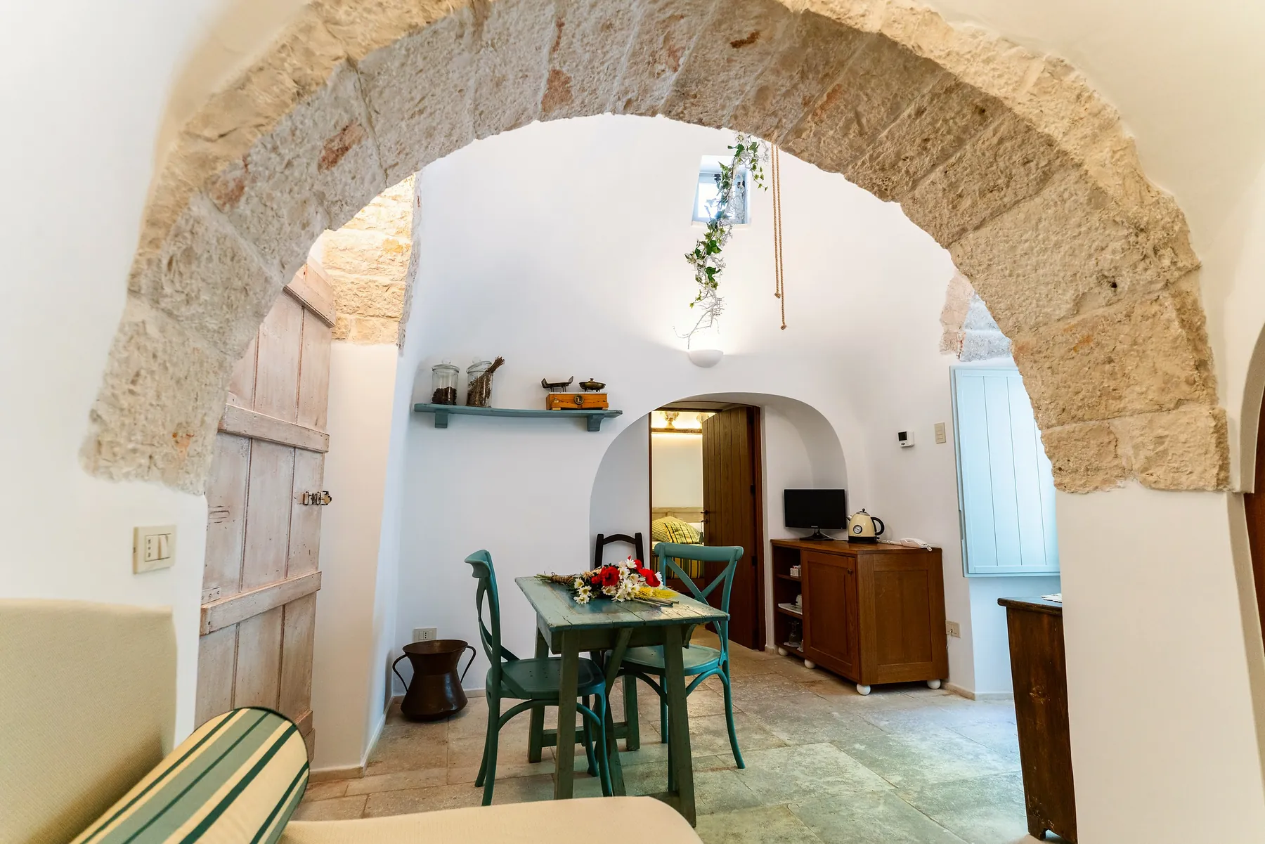 Junior Suite Trullo: a stone arch opening onto a room with an aqua green table with flowers and a wooden dresser.