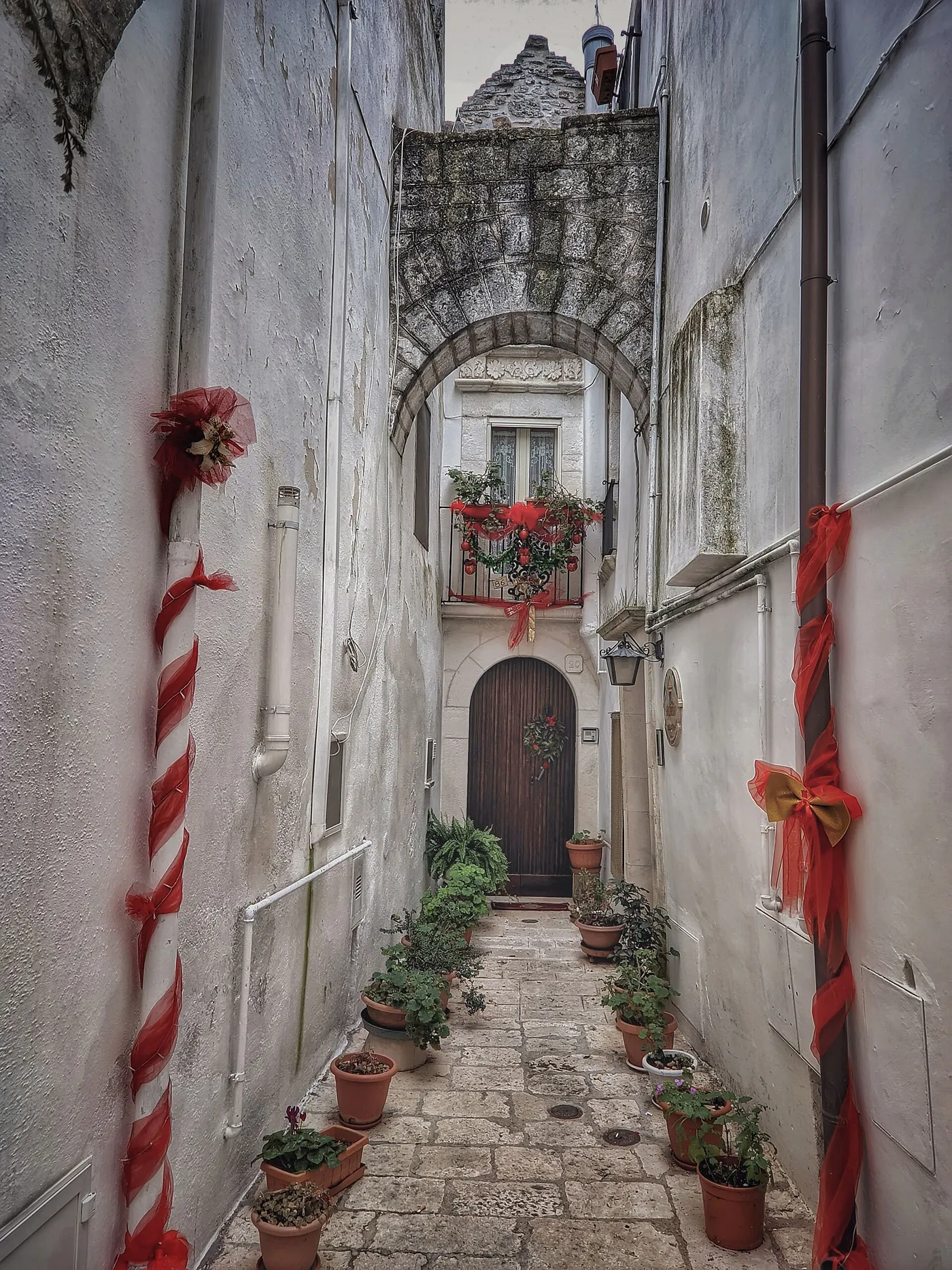 A narrow white lane in Locorotondo under a stone arch, with a red and white striped pole and a pipe wrapped in red tulle with a gold bow, and a garland on the balcony above a wooden door.