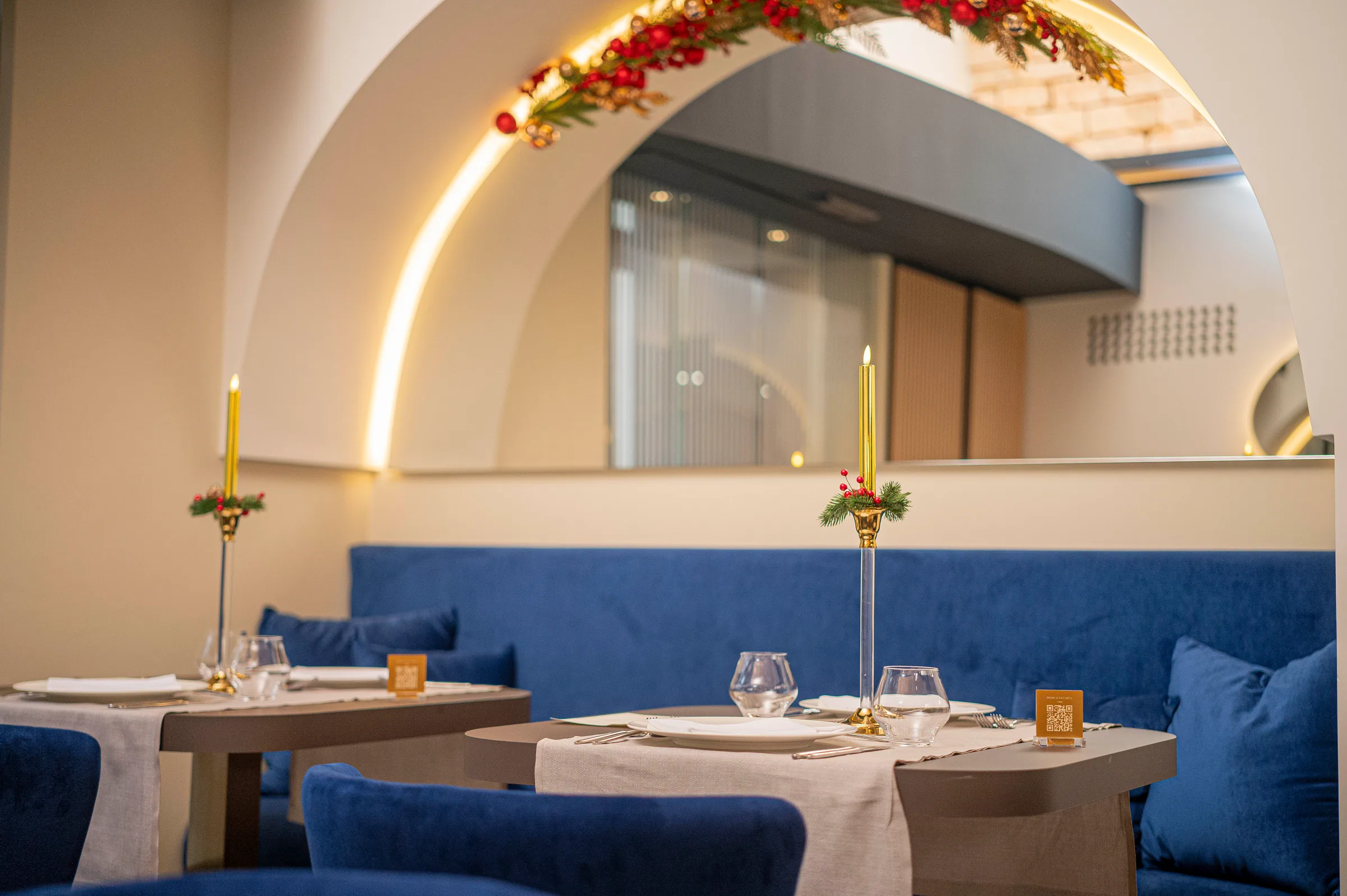 Dining room of NOS restaurant: set tables with gold taper candles and fir sprigs with red berries along a blue velvet bench; a garland of red and gold decorations follows an arched alcove lit by a warm strip light.