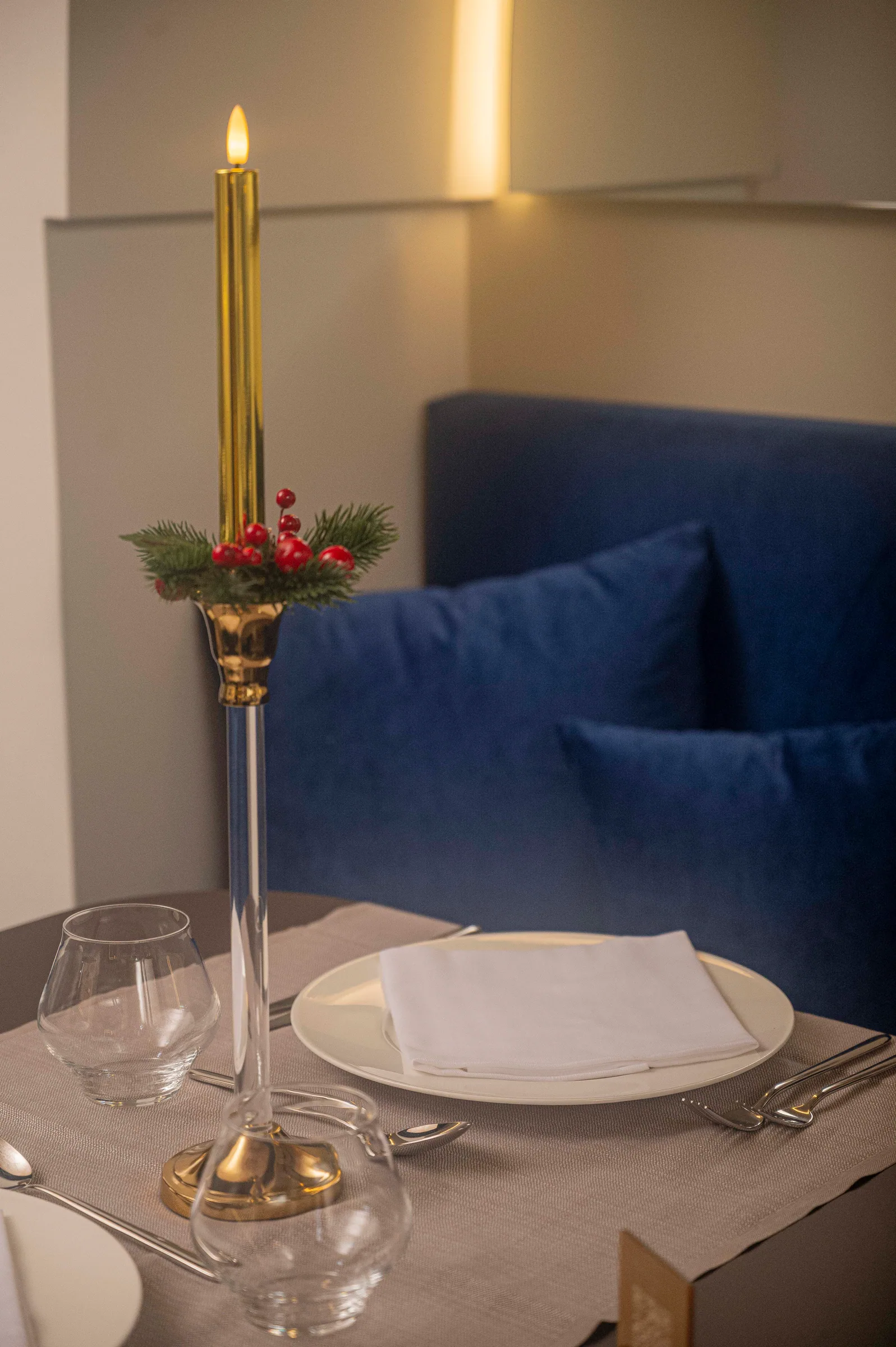 Gold taper candle with a sprig of fir and red berries on a glass candlestick, beside a set place with plate, folded napkin and glasses; a blue velvet bench behind.