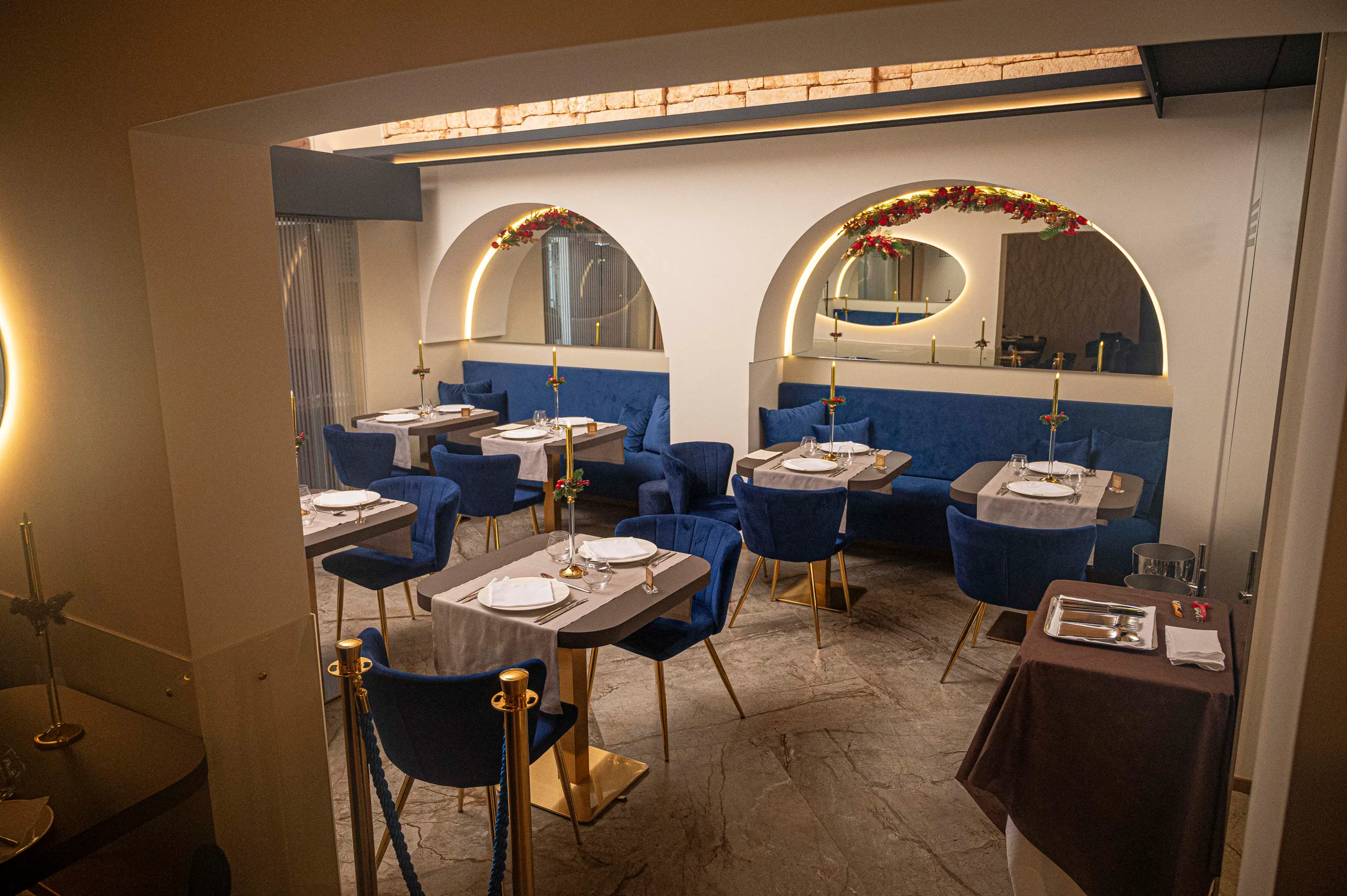 View into the NOS dining room from the entrance: two arched alcoves with garlands, blue benches and set tables; gold posts with a blue rope stand in the foreground.