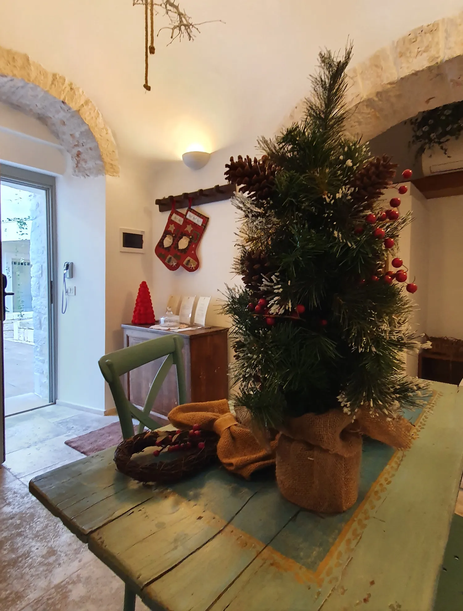 A small Christmas tree in a sackcloth pot on an aqua green wooden table, with a twig wreath; two red stockings hang on a wall rack under white stone arches.