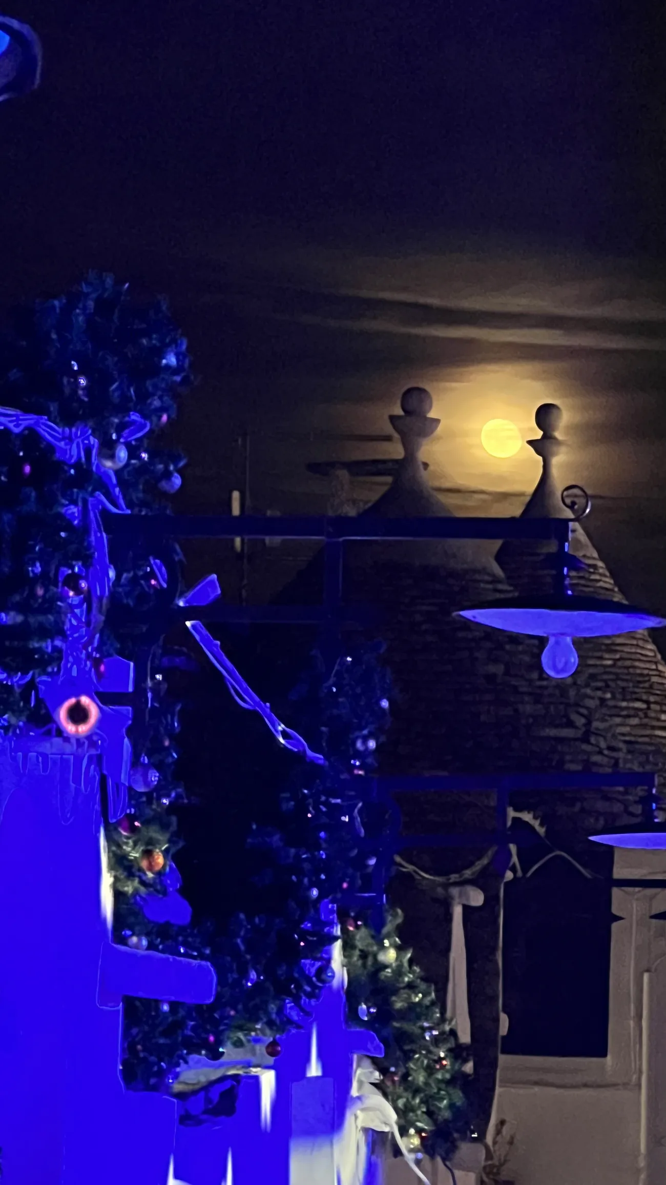 A Christmas garland with baubles and a blue light fixture in front of conical trulli roofs under a full moon, at night in Alberobello.