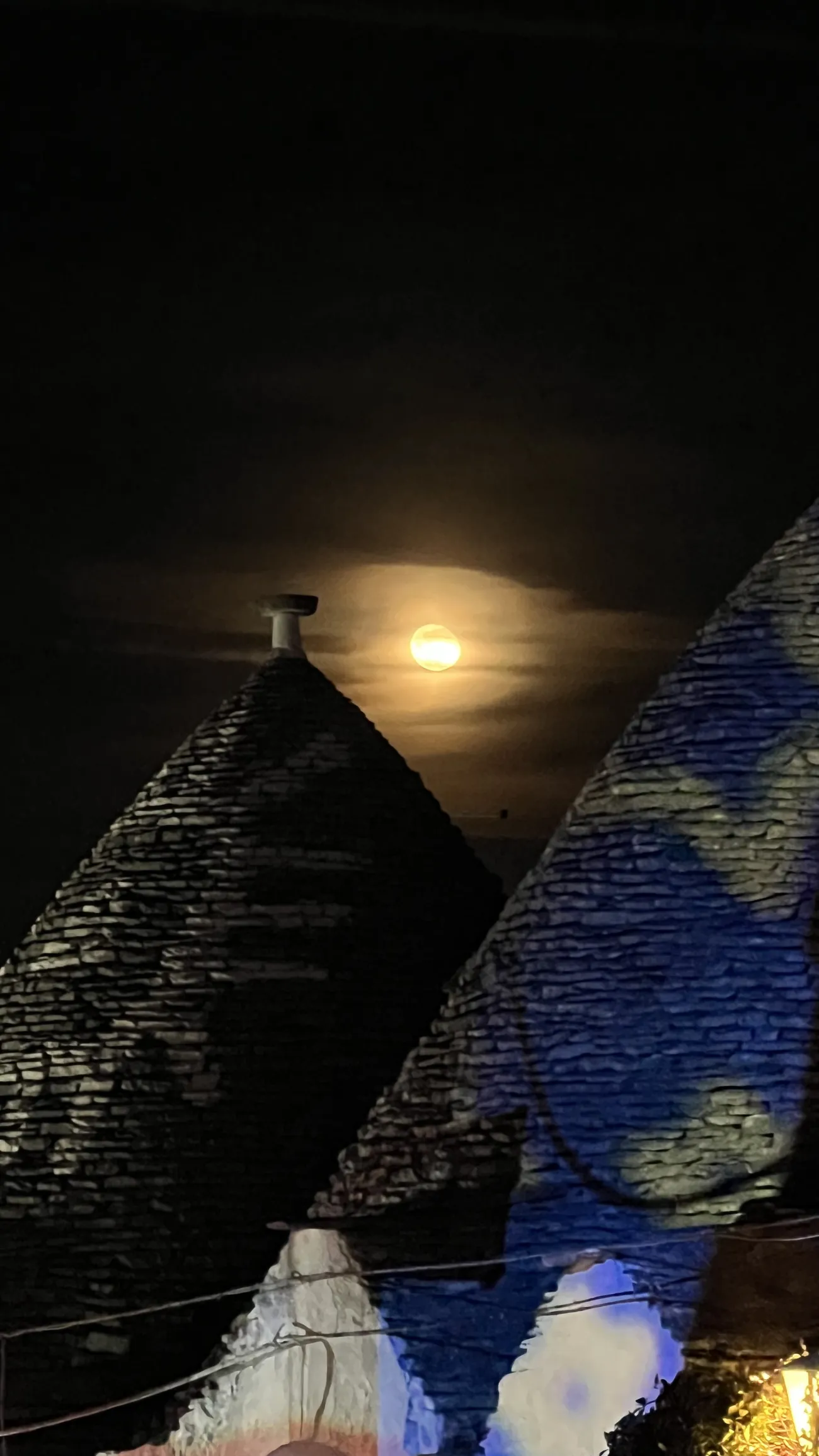 Two conical stone roofs of trulli at night in Alberobello, with the full moon behind one of them.