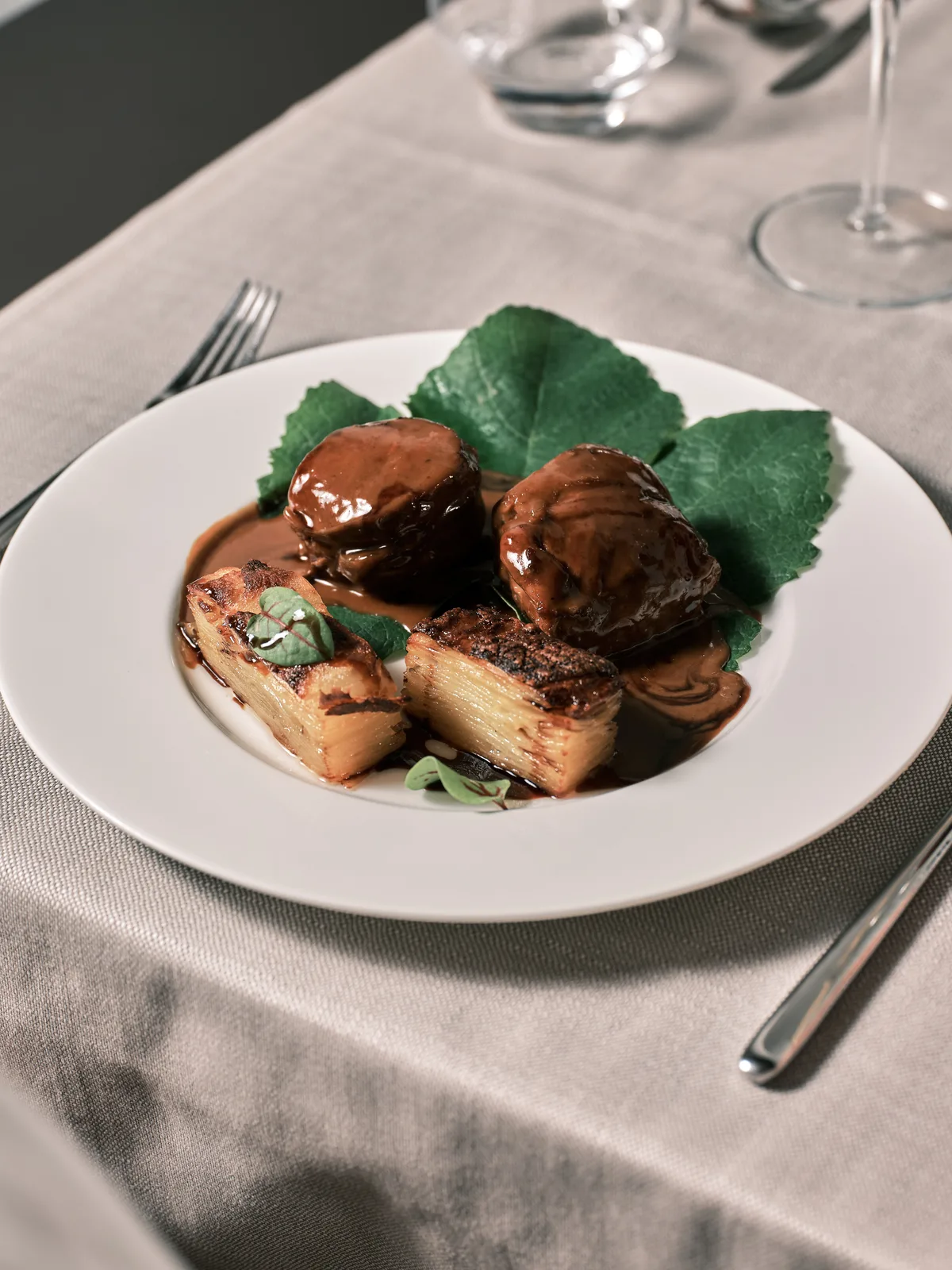A white plate of braised meat with a potato millefeuille and green leaves, on a linen tablecloth with glasses and cutlery.